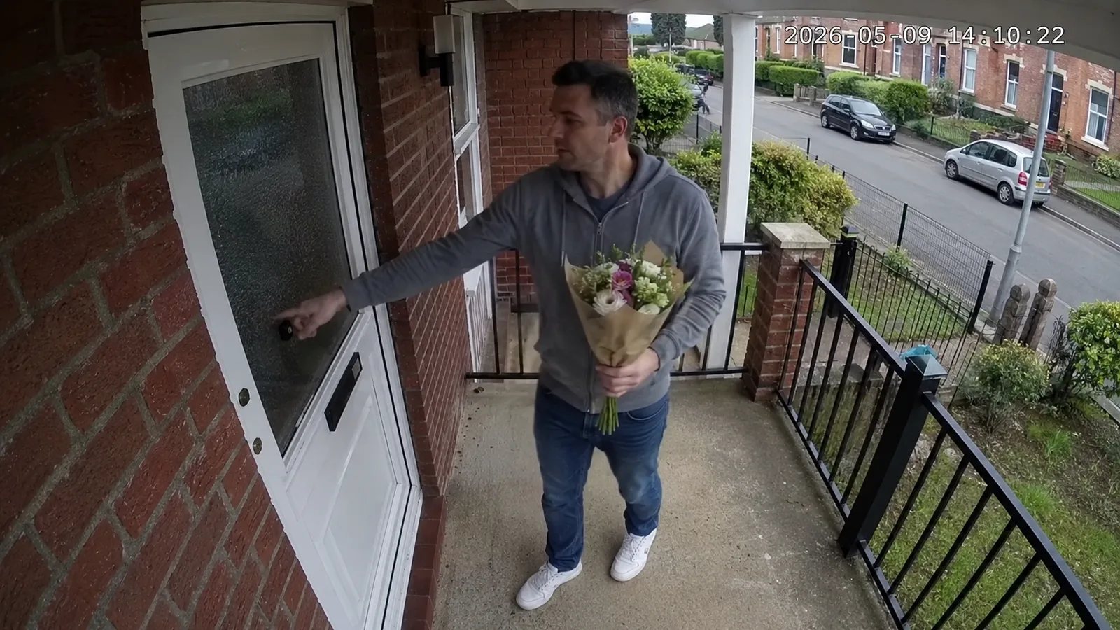 A doorbell-camera still of a man in a grey hoodie at a front door in daylight, holding a bouquet of flowers, timestamp 2026-05-09 14:10:22